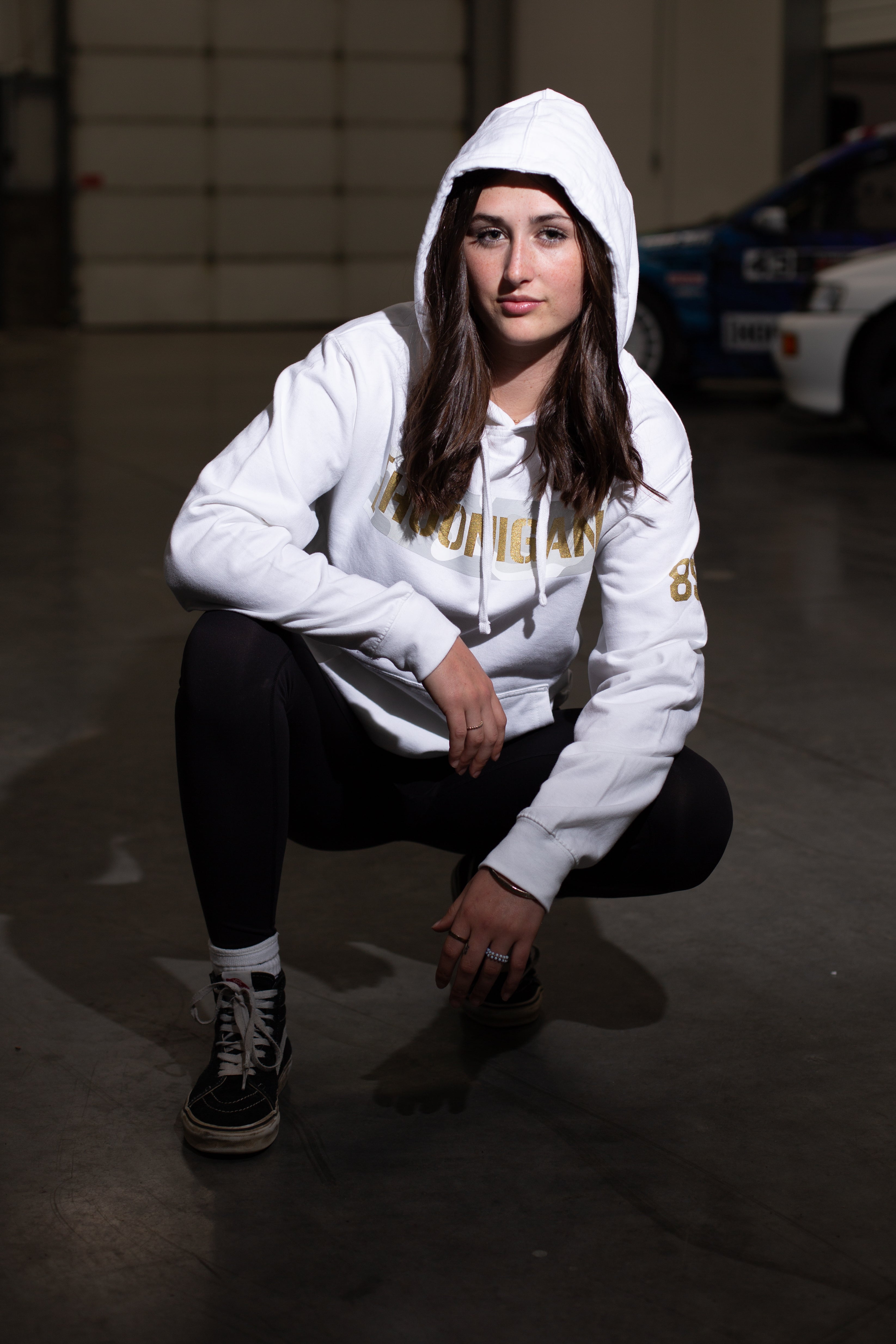 A young woman crouching in a garage wearing a white hoodie with "Snow Camo" and gold lettering, paired with black pants and sneakers.