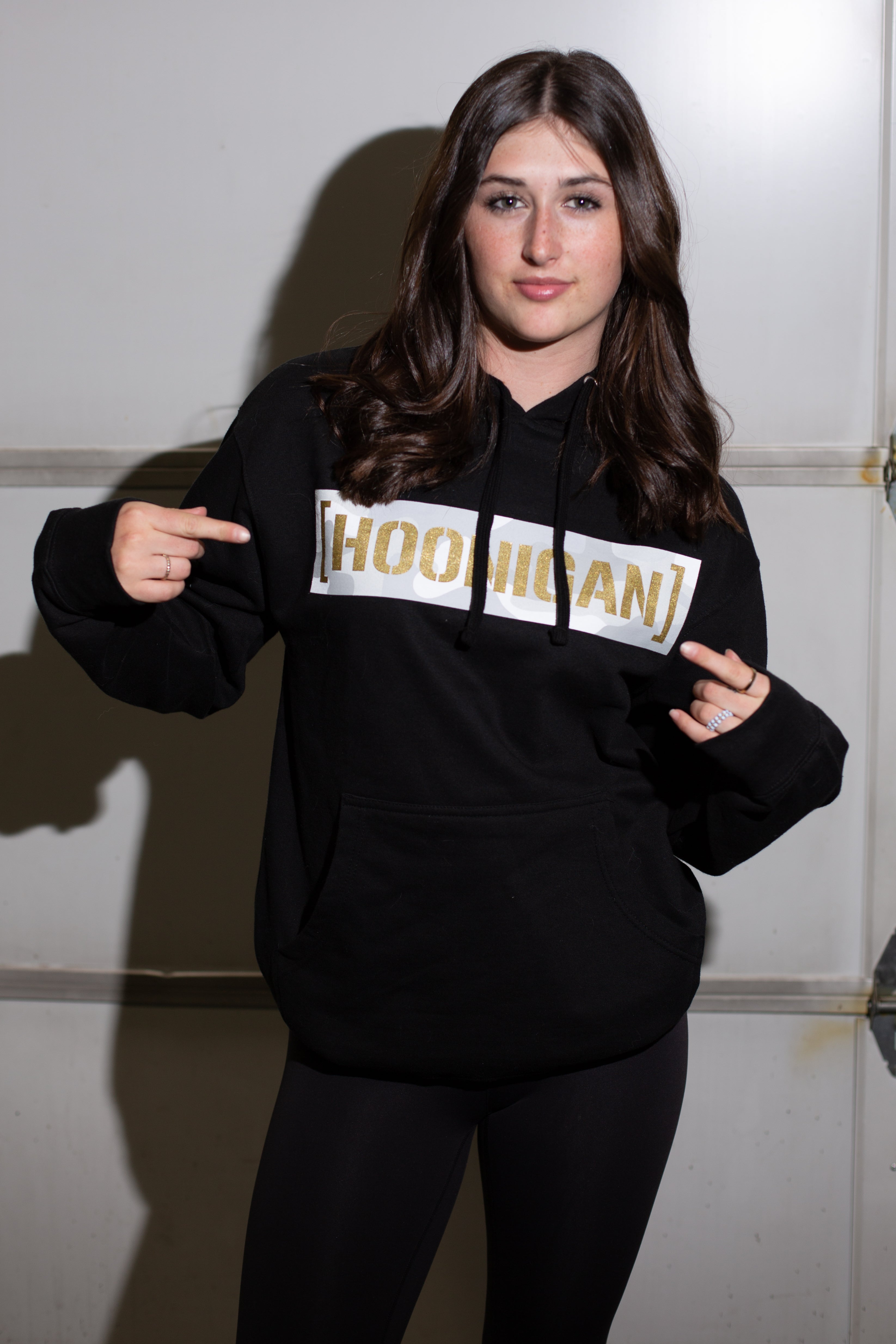 Young woman wearing a black hoodie with "HOODHIGAN" in gold and white text, pointing to the logo on her chest, standing indoors.