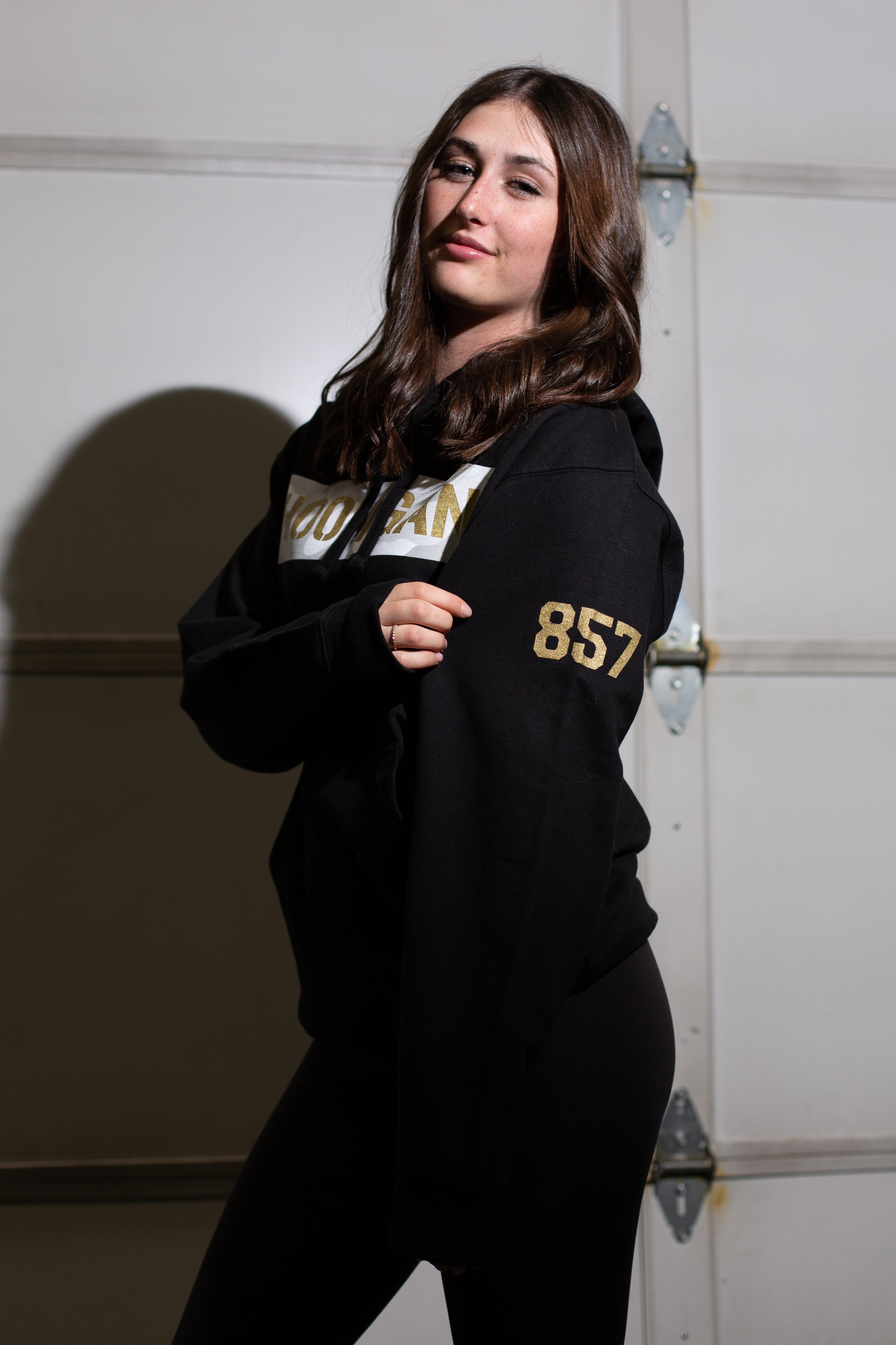 Young woman wearing a black Lia Block SNOW CAMO CENSOR BAR pullover hoodie with gold accents, standing against a garage door.