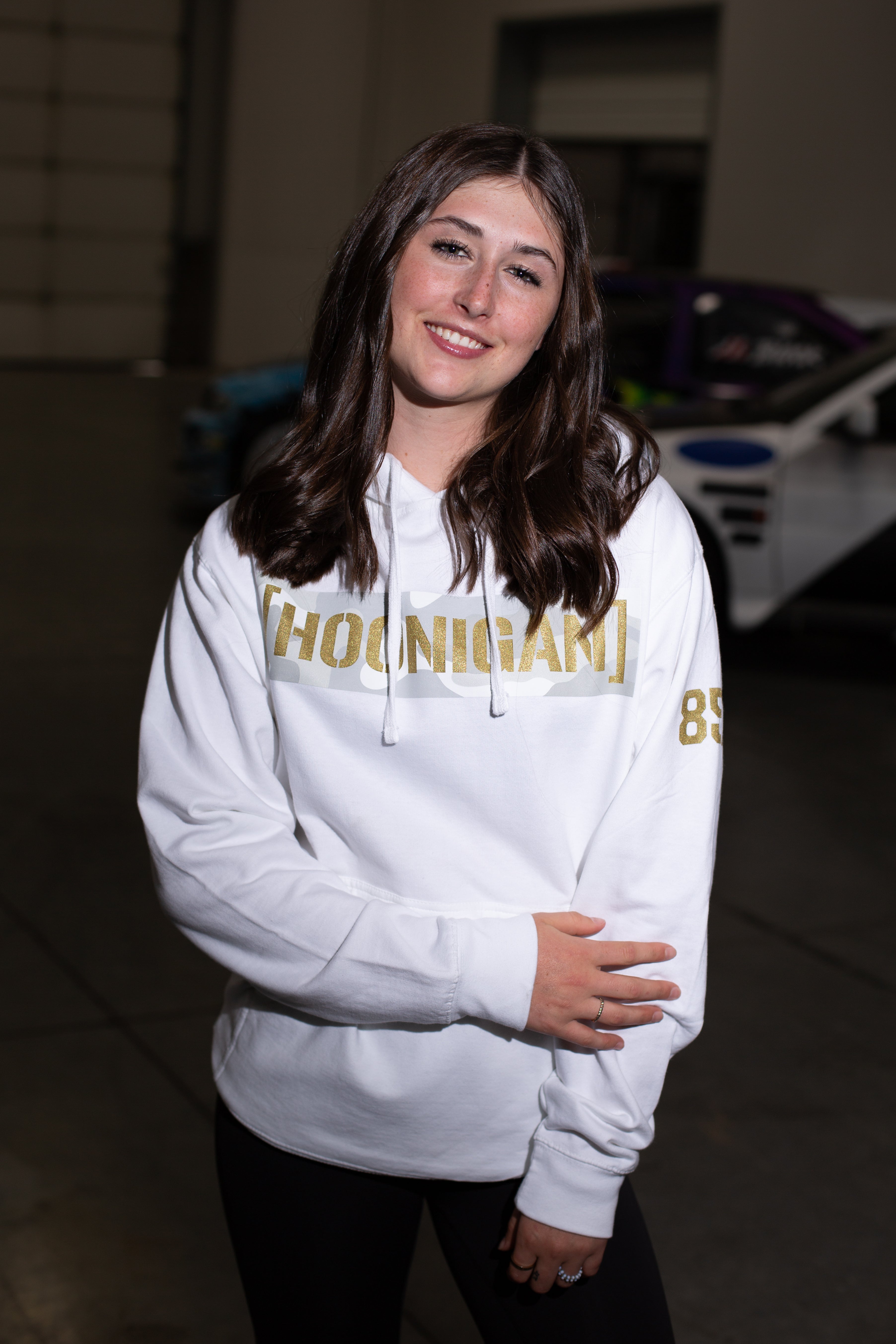 Young woman wearing a white hoodie with "HOONIGAN" and "85" printed in gold, standing indoors with a race car in the background.