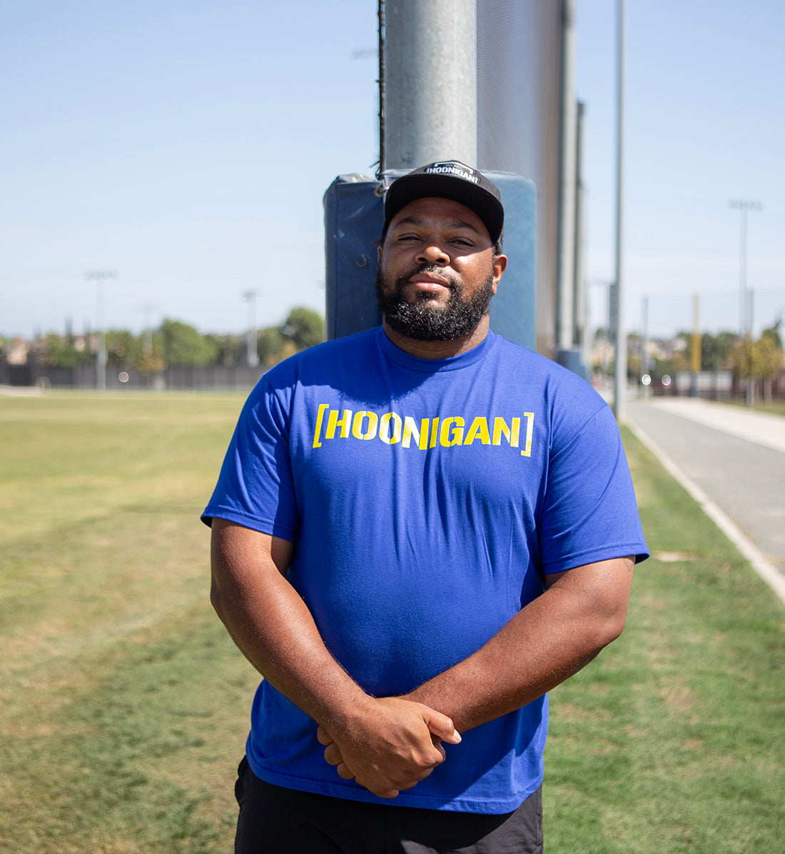 Man wearing a blue Hoonigan T-shirt with yellow logo, standing outdoors near a sports field and a padded post.