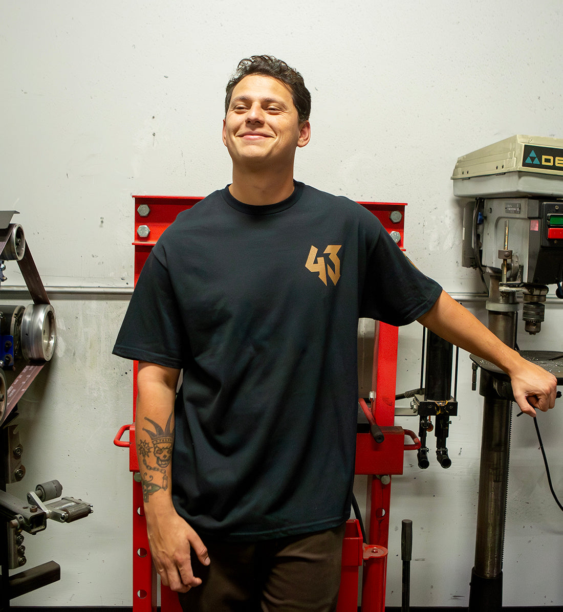 Man wearing a black Hoonigan GYMKHANA7 HITS short sleeve tee, smiling, standing in a workshop with tools and equipment.