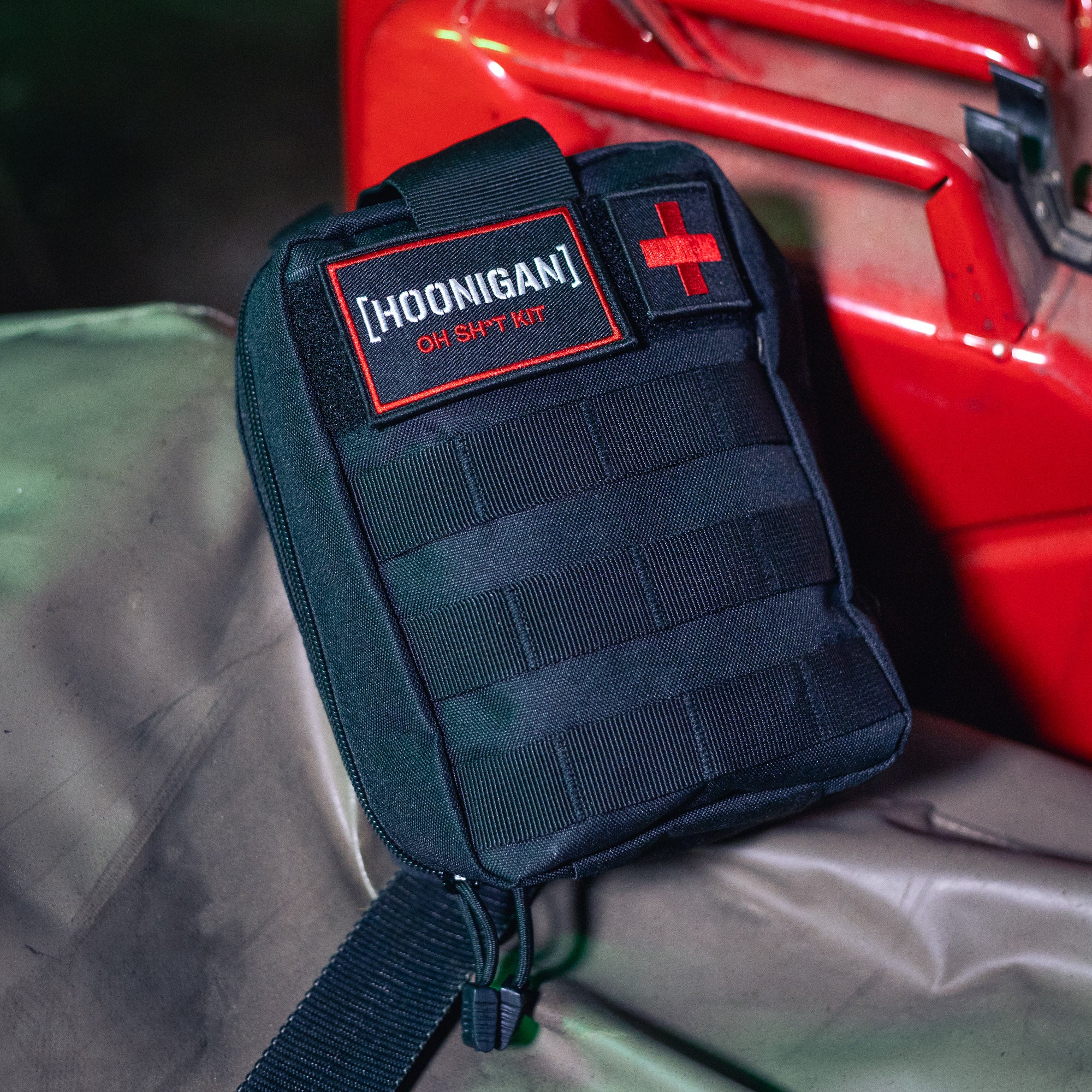 Black Hoonigan OH SH*T Safety Kit pouch with red and white logo, featuring a red cross patch, placed on a surface near a red vehicle.