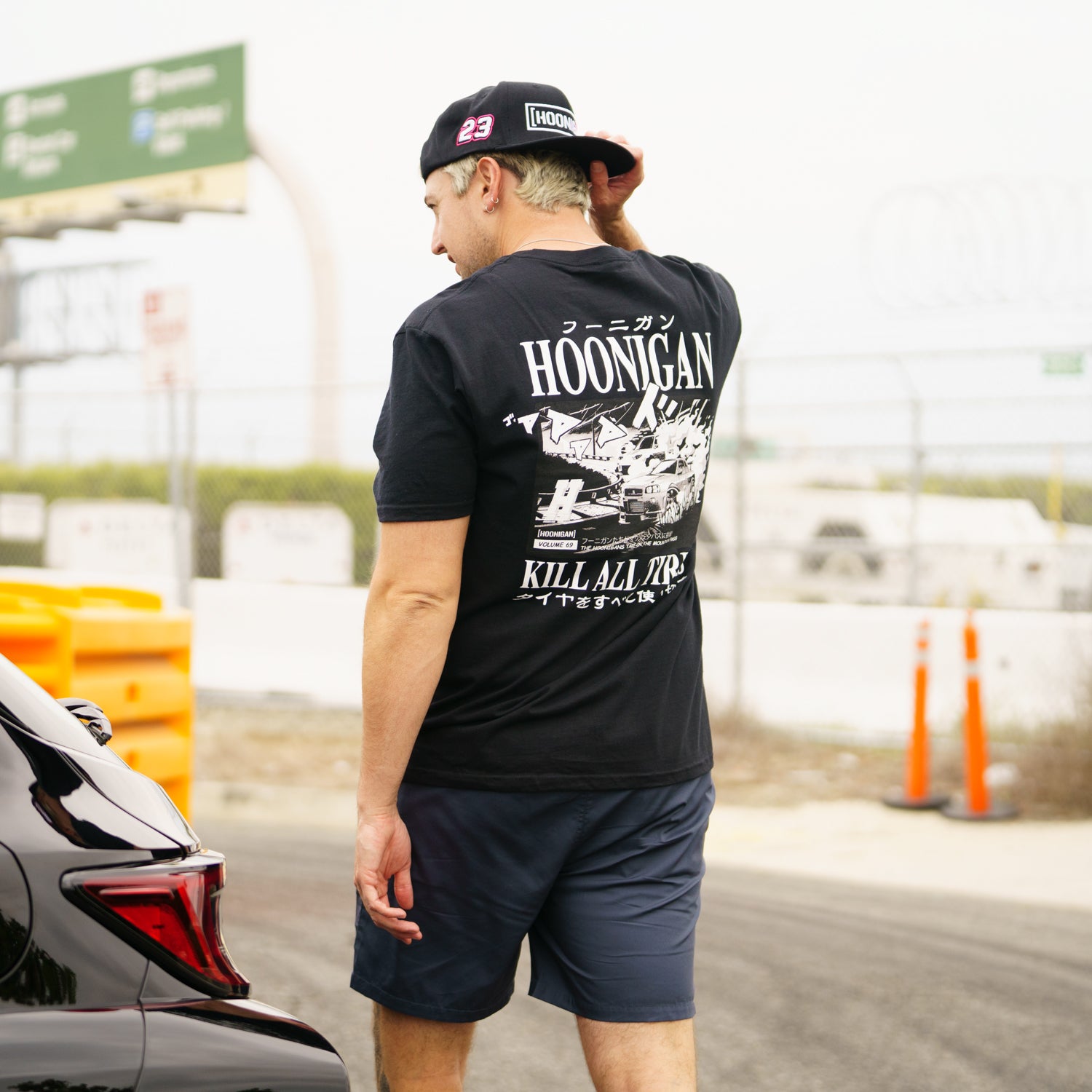 Man wearing a black Hoonigan Tokyo Drip short sleeve tee with graphic print, standing outdoors near a car and orange barriers.