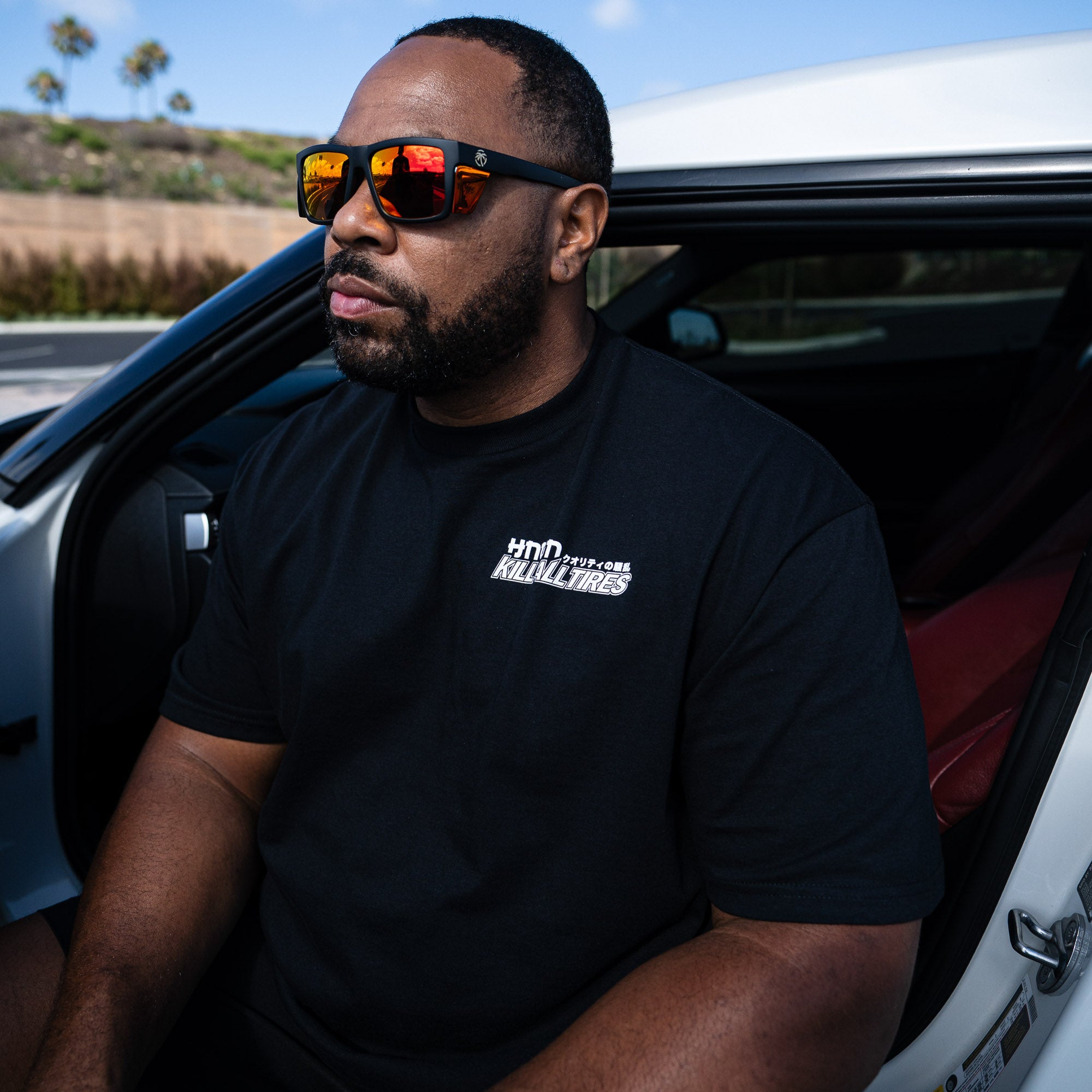 Man wearing a black Hoonigan WANGAN C1 short sleeve tee and sunglasses, sitting in a car with a sporty interior.