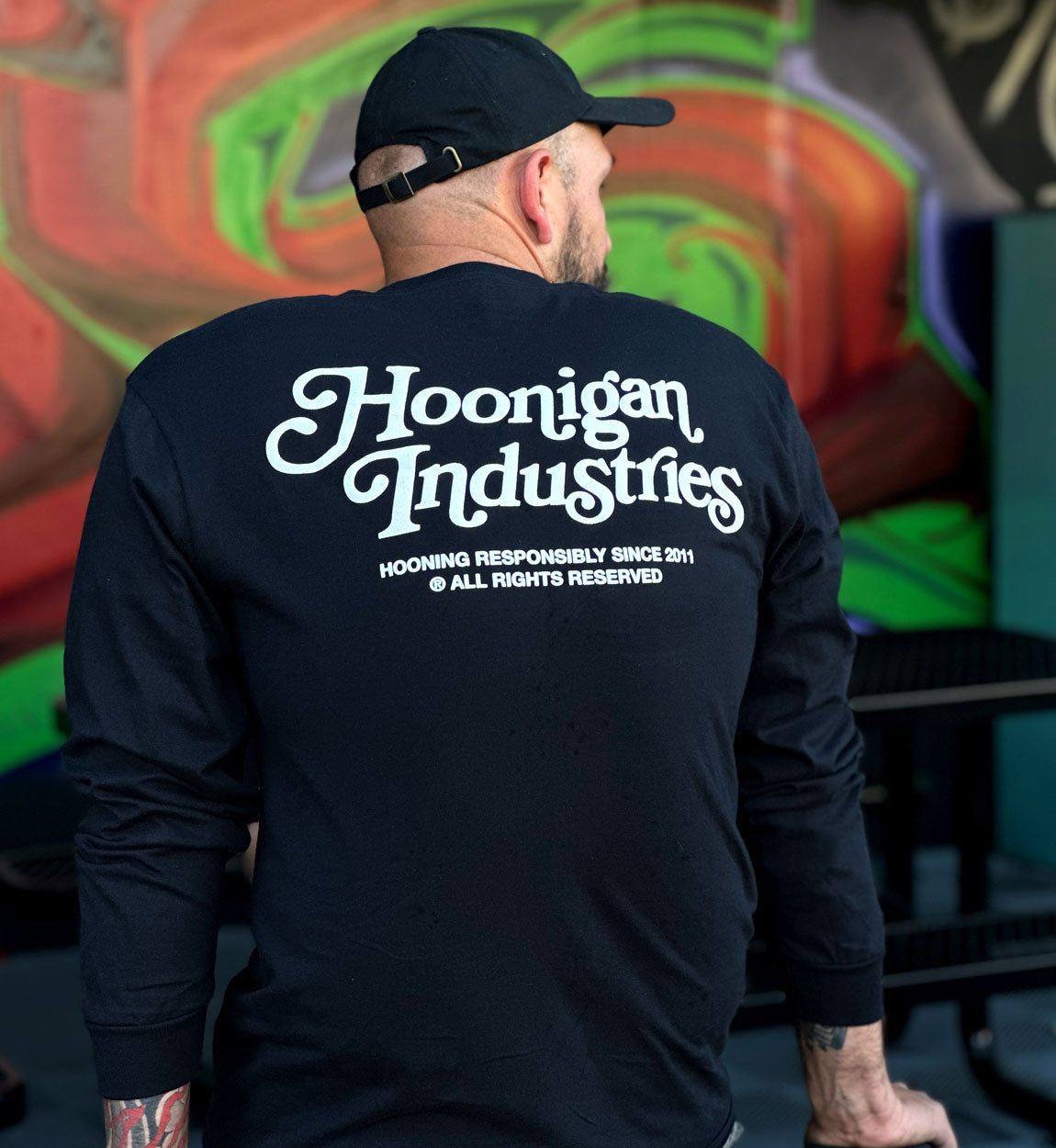 Back view of a man wearing a black long sleeve tee with "Hoonigan Industries" printed on the back, standing in front of a colorful graffiti wall.