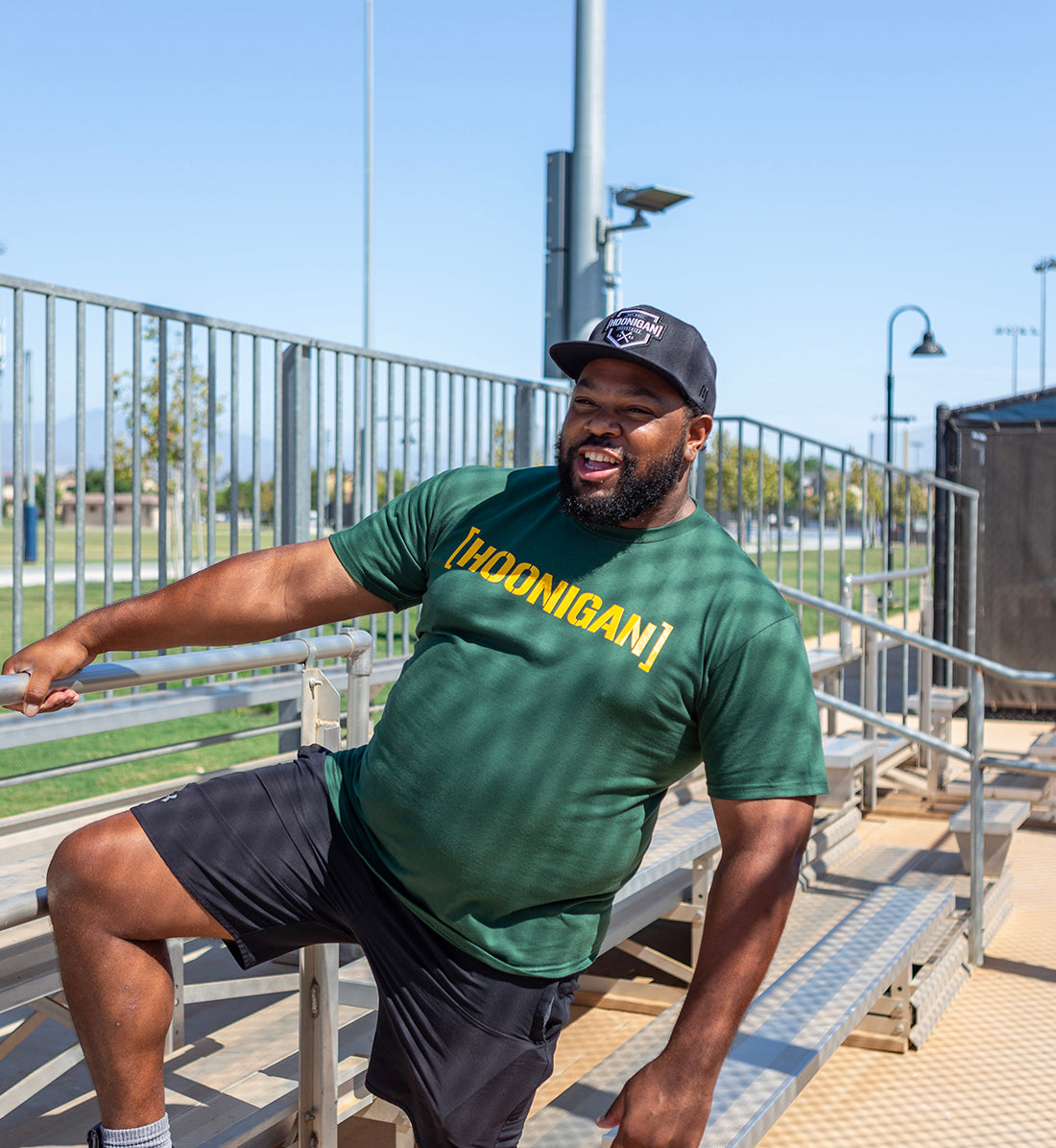 Man wearing a green Hoonigan T-shirt with bold yellow logo, black shorts, and a black cap, sitting on bleachers outdoors.