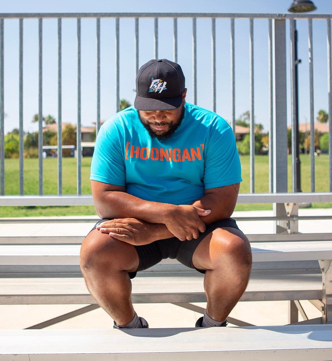 Man sitting on bleachers wearing a blue T-shirt with "[HOONIGAN]" in orange text, black shorts, and a black cap with a logo.