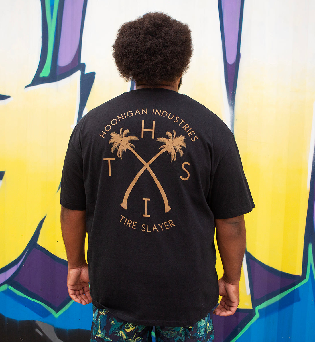 Back view of a person wearing a black Hoonigan GYMKHANA7 HITS short sleeve tee with palm tree graphics and text, standing in front of colorful graffiti.