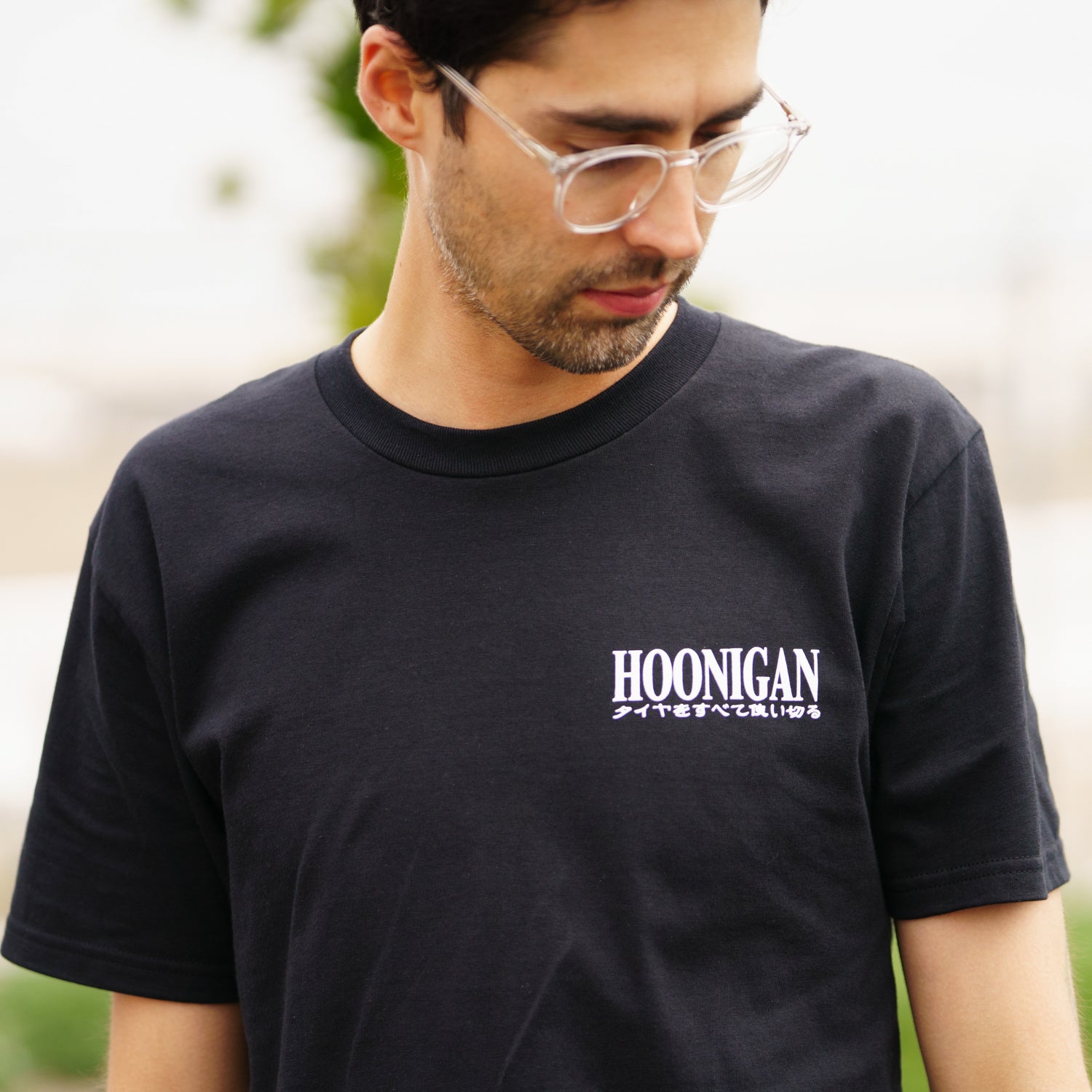 Close-up of a man wearing a black Hoonigan TOKYO DRIP short sleeve T-shirt with white logo and Japanese text, outdoors.
