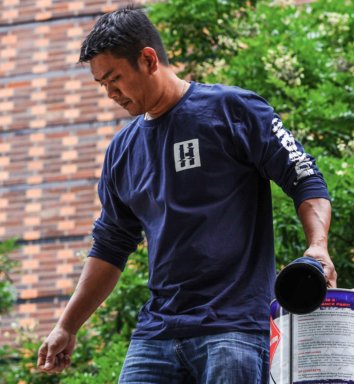 Male model wearing a navy long sleeve tee with "GLITCH BRACKET" graphic on the front and sleeve, outdoors with greenery.